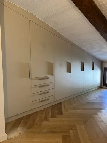 Long linéaire de placards beiges sous pente avec portes battantes et tiroirs, dans un couloir mansardé au parquet chevrons.