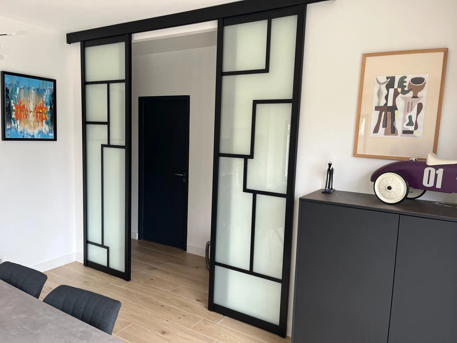 Double porte coulissante suspendue à cadre noir et panneaux de verre dépoli, séparant séjour et couloir.